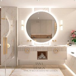 Anna Design by Anna Stenka-Kosińska - Elegancka łazienka z okrągłym lustrem podświetlanym LED, beżowymi płytkami i złotymi akcentami, w tym armaturą i uchwytami szafek. Widoczna kabina prysznicowa i kwiaty.