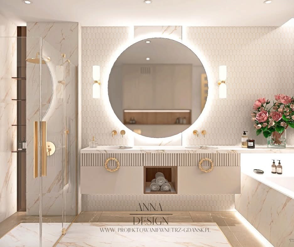 Elegancka łazienka z okrągłym lustrem podświetlanym LED, beżowymi płytkami i złotymi akcentami, w tym armaturą i uchwytami szafek. Widoczna kabina prysznicowa i kwiaty.