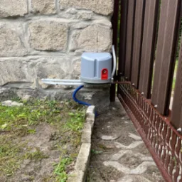 Szary napęd bramy wjazdowej zamontowany na kamiennym słupku, z widoczną brązową bramą z dekoracyjnymi elementami. U dołu kostka brukowa i trawa.