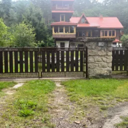 Ciemnobrązowa brama wjazdowa z ozdobnymi elementami, osadzona między kamiennymi słupkami. W tle widoczny dom z czerwonym dachem i bujna zieleń.