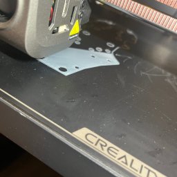 Patryk Kołcon - Drukarka 3D Creality w trakcie pracy: szary element z otworami powstaje na czarnym stole roboczym. Widoczna głowica drukująca i fragment napisu CREALITY.