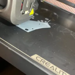 Drukarka 3D Creality w trakcie pracy: szary element z otworami powstaje na czarnym stole roboczym. Widoczna głowica drukująca i fragment napisu CREALITY.