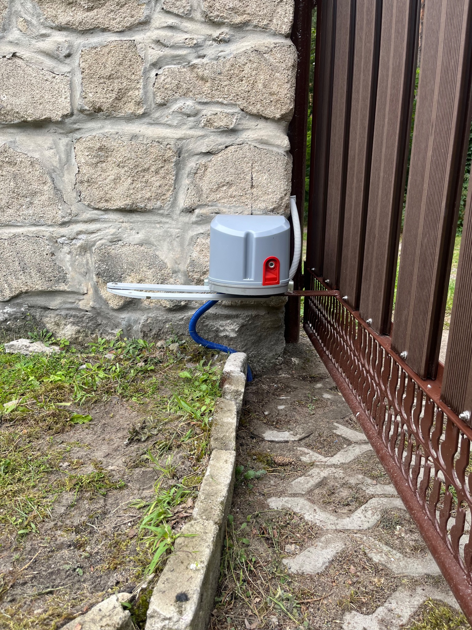 Szary napęd bramy wjazdowej zamontowany na kamiennym słupku, z widoczną brązową bramą z dekoracyjnymi elementami. U dołu kostka brukowa i trawa.