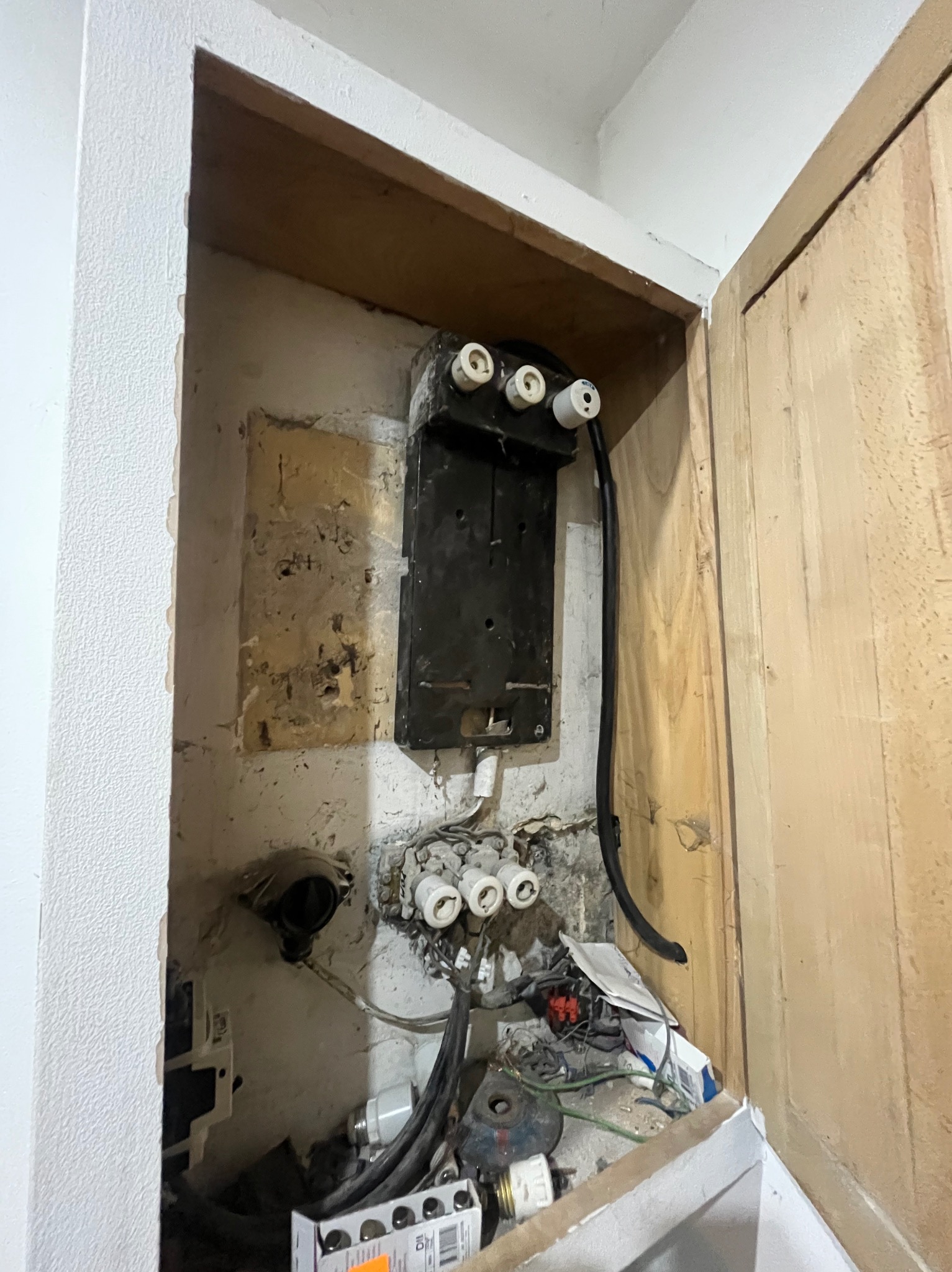 Stara, czarna skrzynka elektryczna z ceramicznymi bezpiecznikami wnęce. Widoczne kable, fragment ściany z odrapaną farbą i drewniane drzwi szafki.