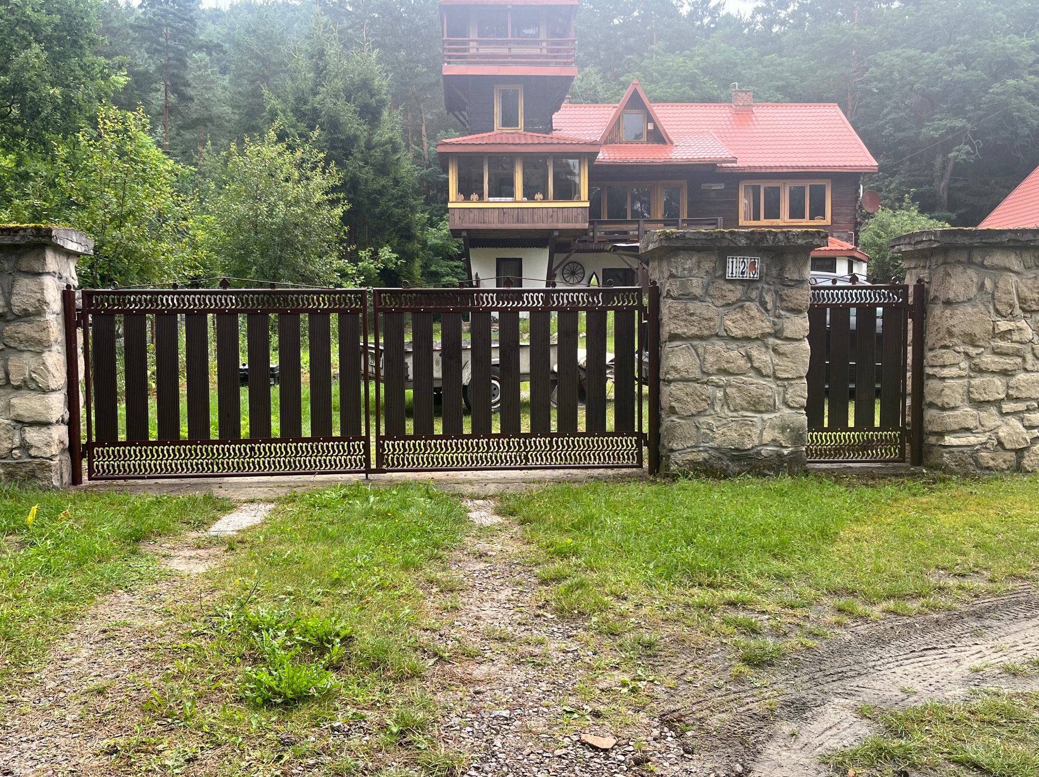 Ciemnobrązowa brama wjazdowa z ozdobnymi elementami, osadzona między kamiennymi słupkami. W tle widoczny dom z czerwonym dachem i bujna zieleń.