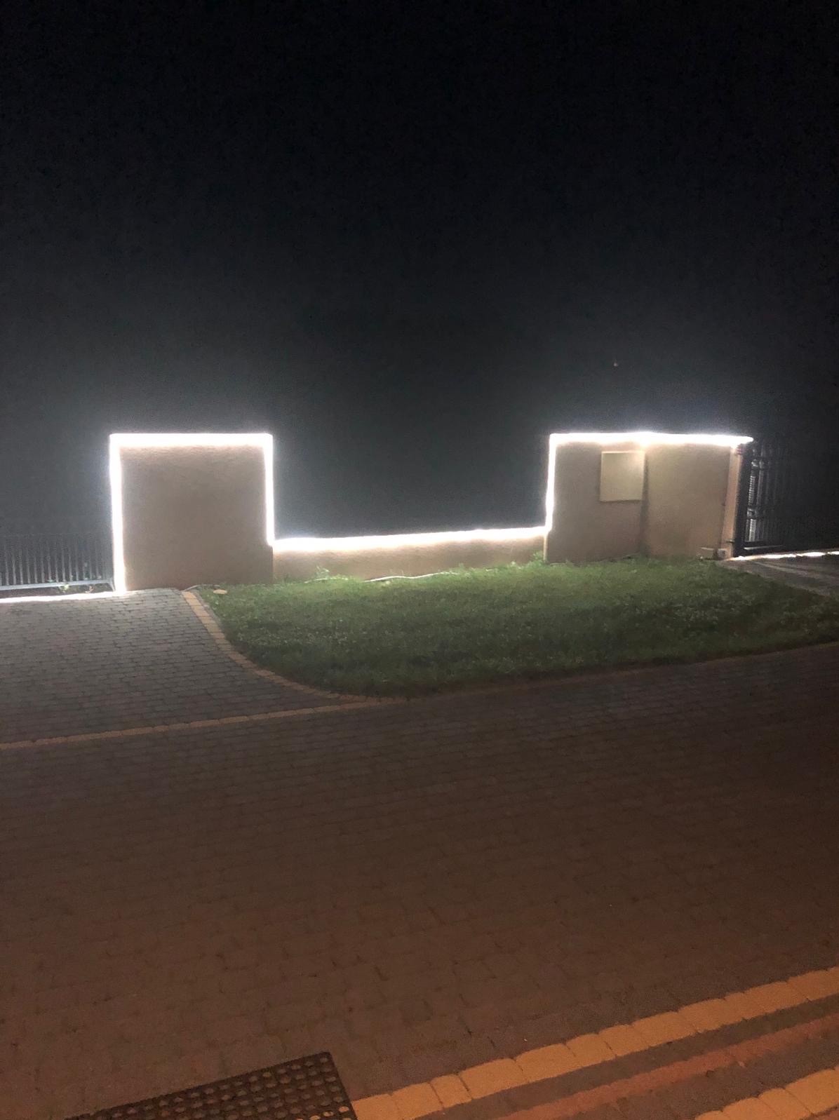 Nocne ujęcie podświetlonych ledami słupków i murku przy bramie wjazdowej. Widoczny fragment chodnika z kostki brukowej i trawnik. Oświetlenie zewnętrzne.