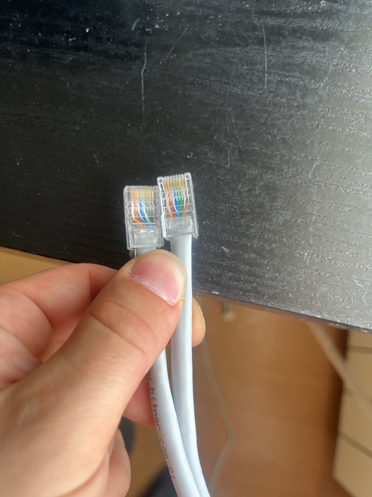 Dwa białe kable Ethernet z przezroczystymi wtyczkami RJ45, trzymane w dłoni, na tle ciemnego blatu. Widoczne kolorowe przewody wewnątrz wtyczek.