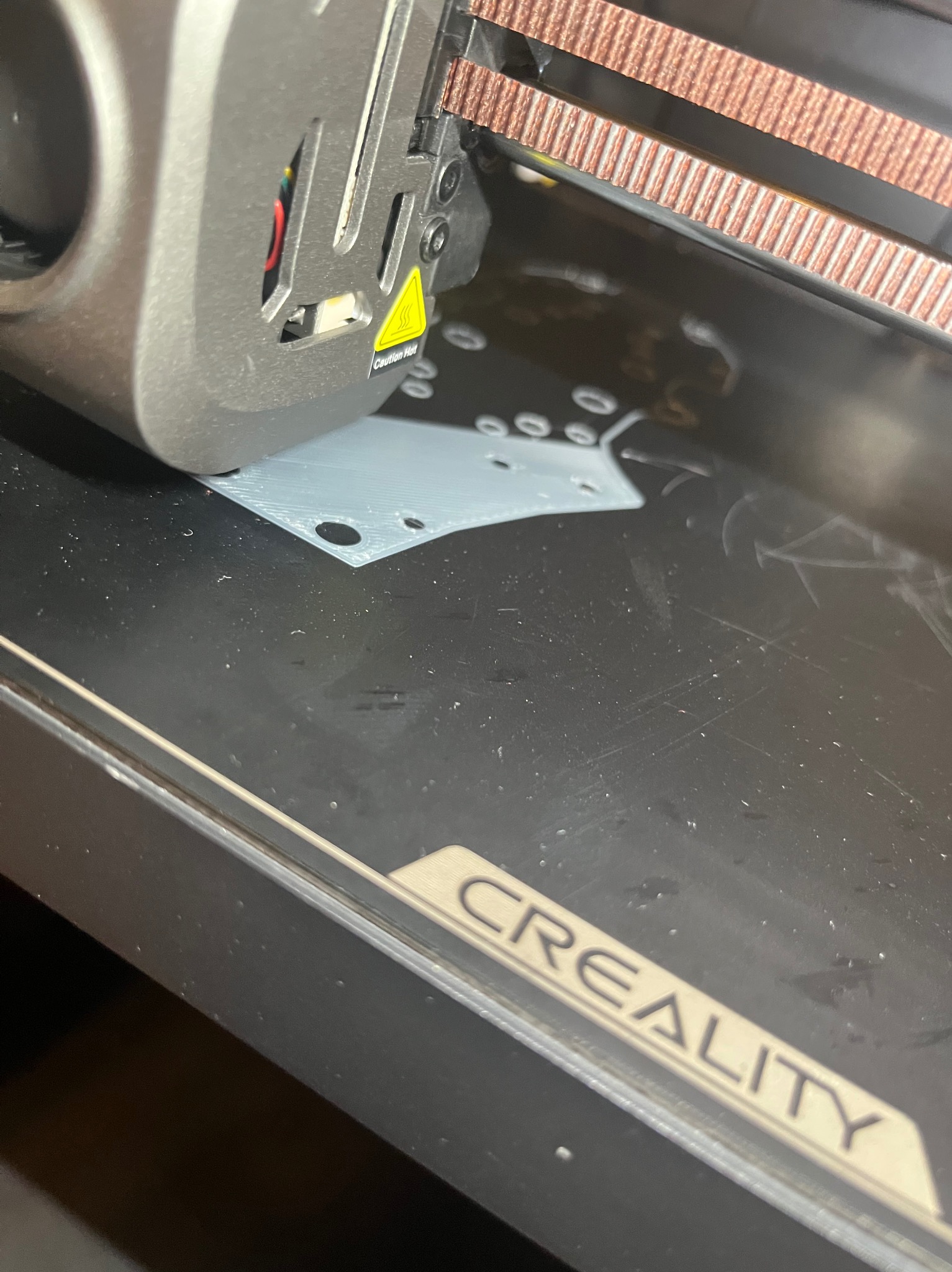 Drukarka 3D Creality w trakcie pracy: szary element z otworami powstaje na czarnym stole roboczym. Widoczna głowica drukująca i fragment napisu CREALITY.