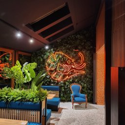 Dariusz Byliniak - Stylowe wnętrze kawiarni z neonem w kształcie słonia na ścianie z zieleni, błękitnymi kanapami i fotelem. Nowoczesny design z elementami roślinnymi i mozaiką.