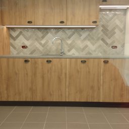 Dariusz Byliniak - Nowoczesna zabudowa kuchenna z drewnianymi frontami, szarą mozaiką na ścianie i subtelnym oświetleniem LED, widok na blat z umywalką i baterią.