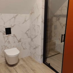 Dariusz Byliniak - Wykończona łazienka na poddaszu: białe marmurowe płytki, podwieszana toaleta z czarnym przyciskiem, kabina prysznicowa z czarną ramą i drewnopodobna podłoga.