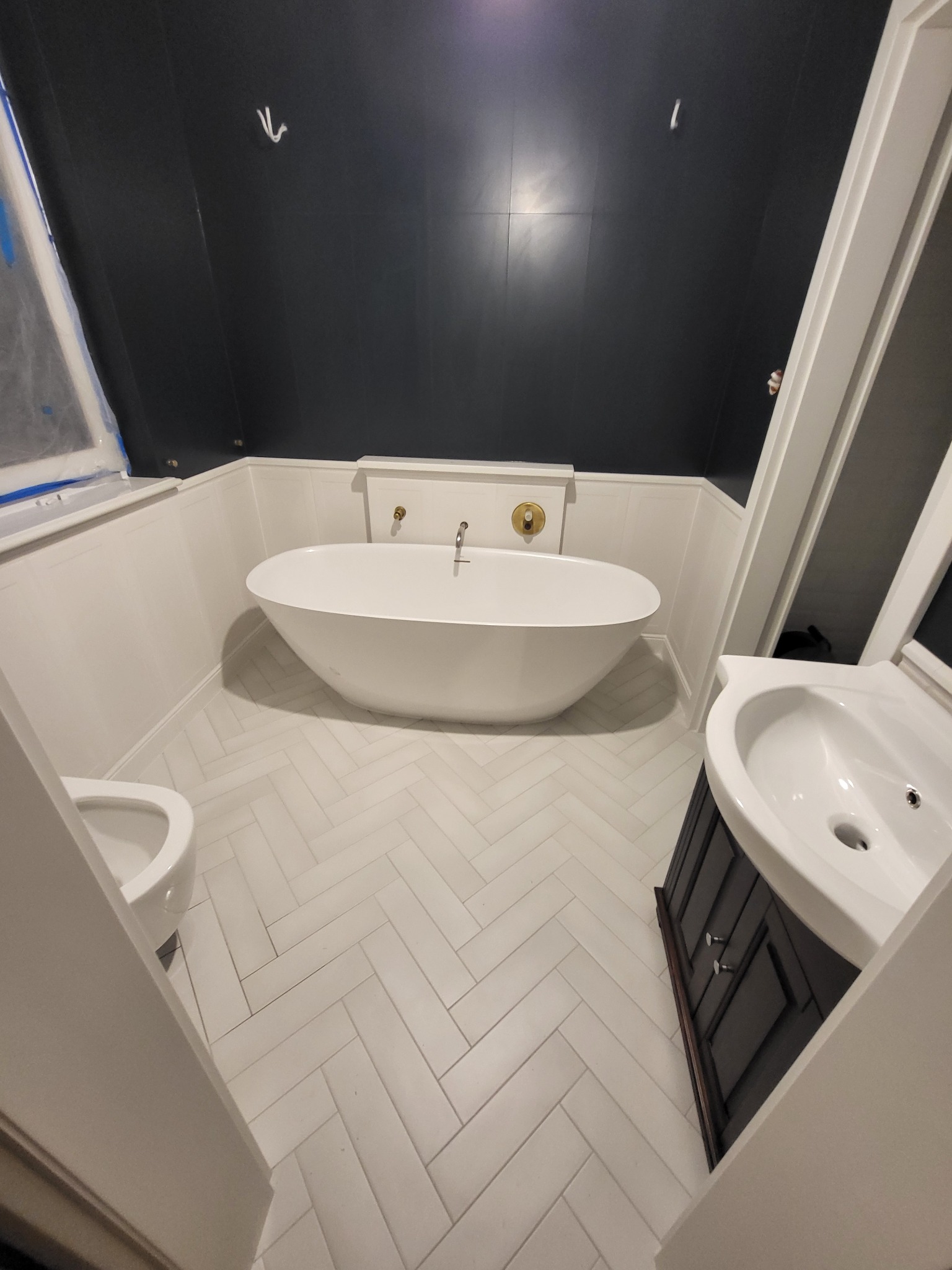 Nowoczesna łazienka z wolnostojącą wanną, umywalką z szafką i toaletą. Ciemne ściany kontrastują z białą armaturą i podłogą w jodełkę.