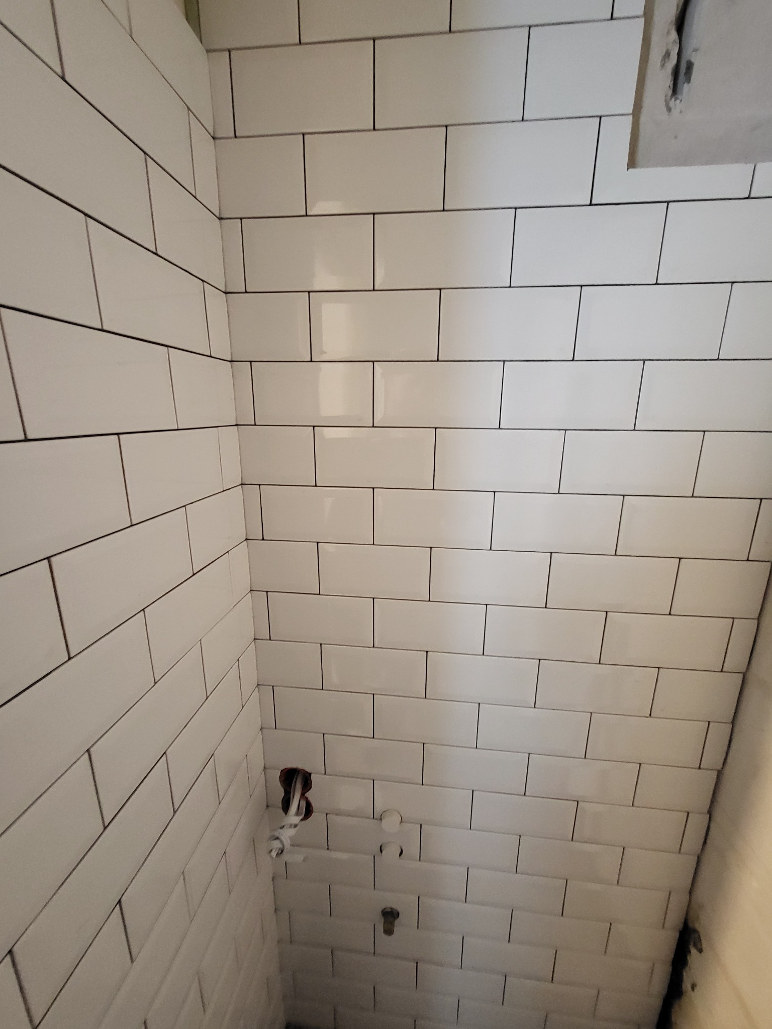 Białe, prostokątne płytki z ciemną fugą, ułożone na ścianach w łazience. Widoczne wystające rury i otwór na instalację elektryczną.