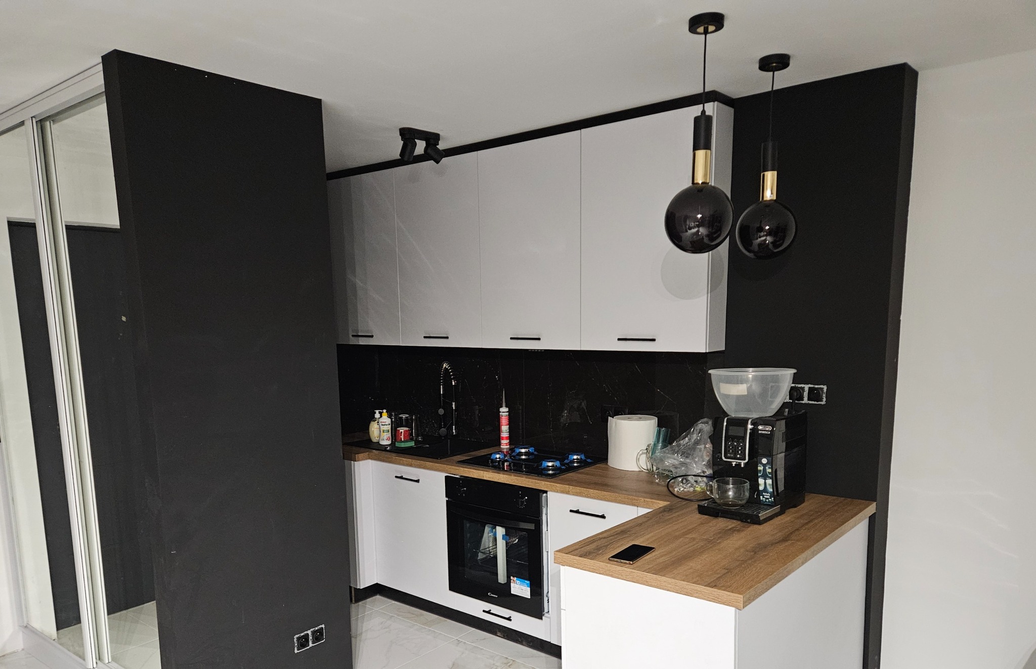 Nowoczesna kuchnia z białymi szafkami, czarnym blatem i ścianą. Dwie czarne lampy wiszące dodają elegancji. Widoczny ekspres do kawy i przesuwne drzwi.