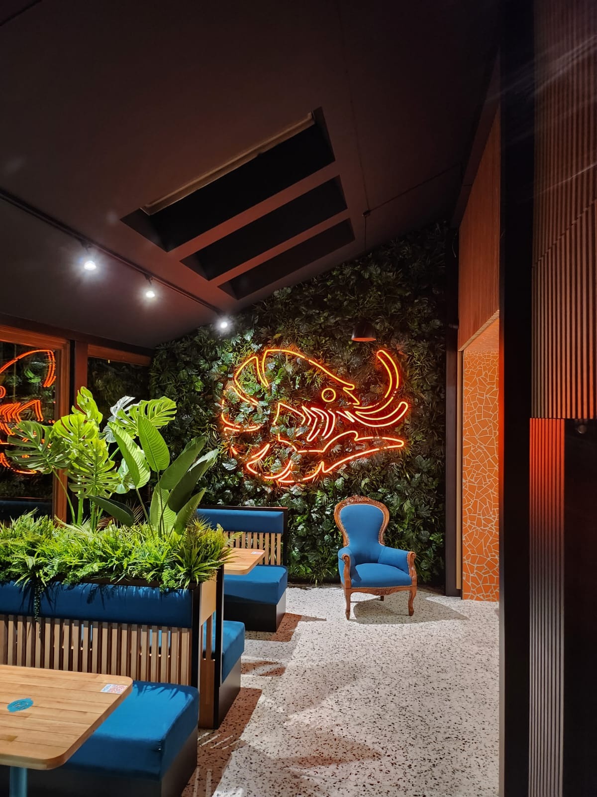 Stylowe wnętrze kawiarni z neonem w kształcie słonia na ścianie z zieleni, błękitnymi kanapami i fotelem. Nowoczesny design z elementami roślinnymi i mozaiką.