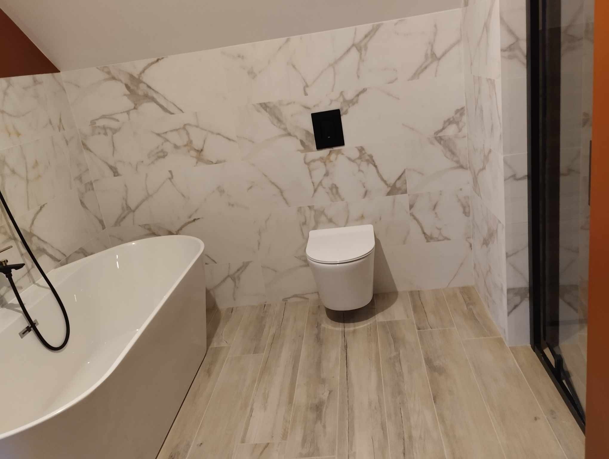 Nowoczesna łazienka z wolnostojącą wanną, podwieszaną toaletą i marmurowymi płytkami ściennymi. Podłoga wyłożona płytkami imitującymi drewno.