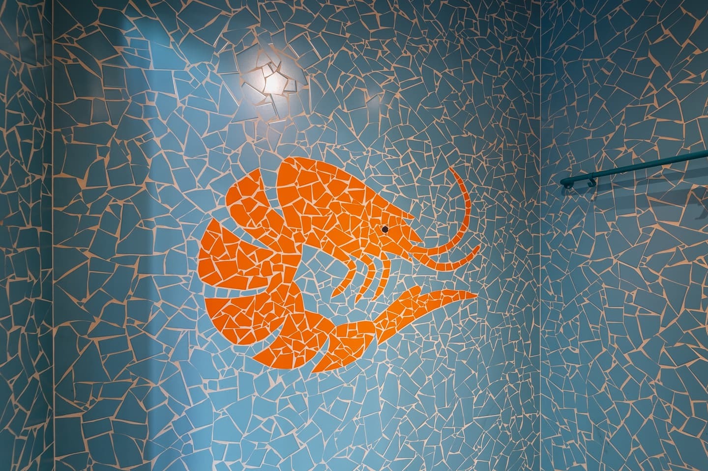 Niebieska mozaika ścienna z pomarańczowym motywem krewetki w łazience. Fragment poręczy w tle. Unikalna aranżacja z detalami.