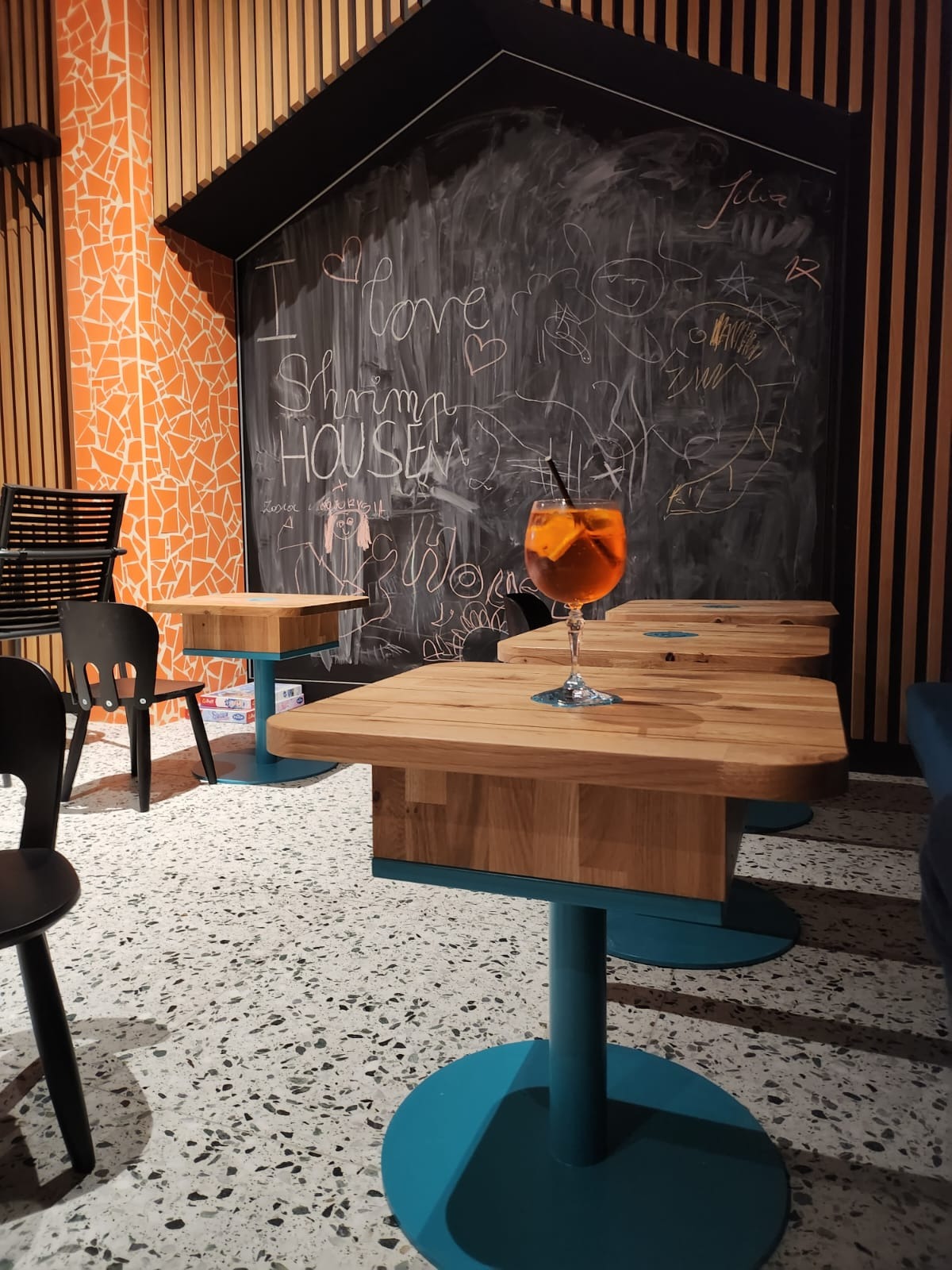 Wnętrze lokalu: drewniane stoliki na turkusowych nogach, czarne krzesła, ściana z pomarańczowej mozaiki i tablica kredowa z rysunkami.