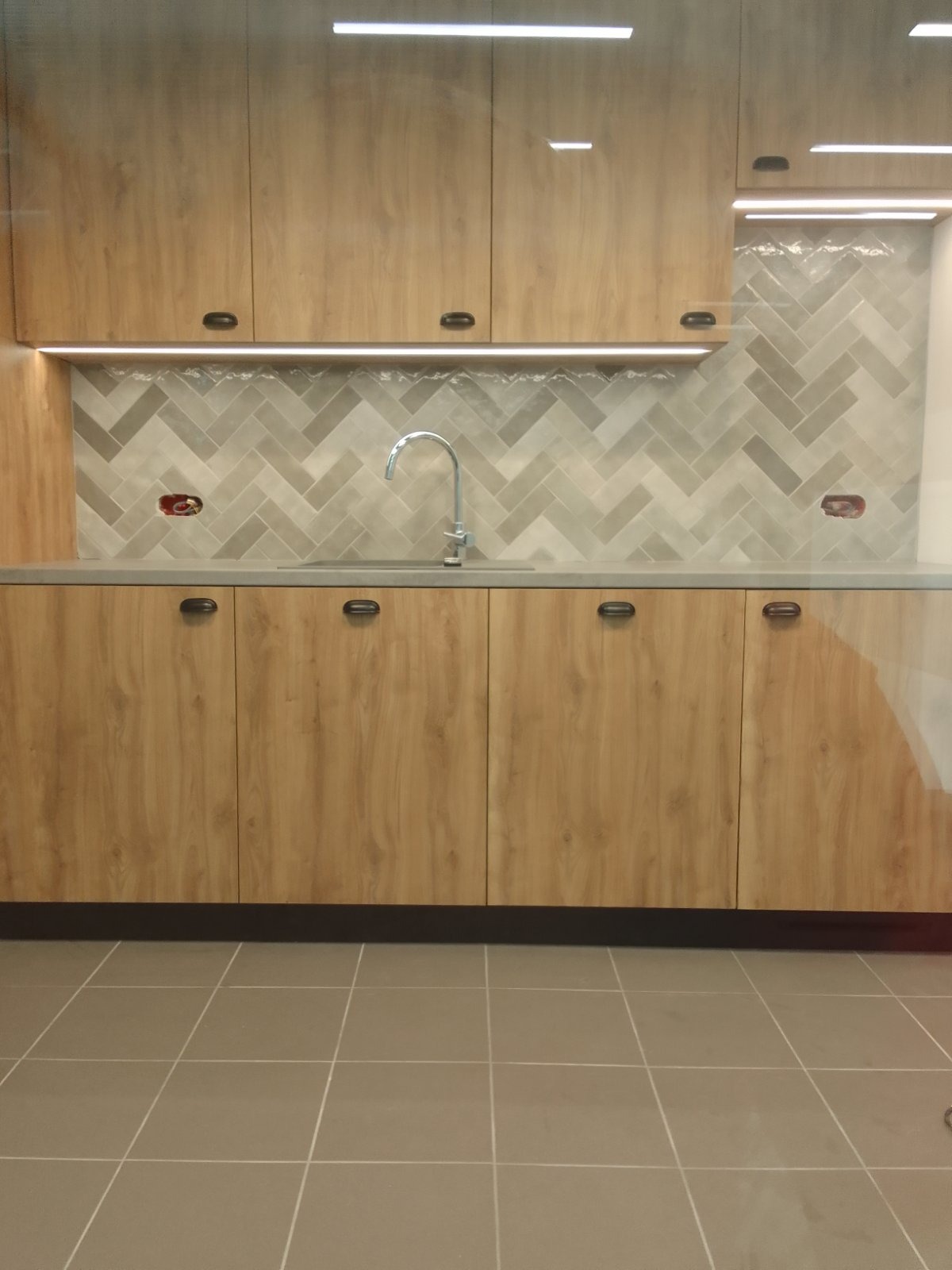 Nowoczesna zabudowa kuchenna z drewnianymi frontami, szarą mozaiką na ścianie i subtelnym oświetleniem LED, widok na blat z umywalką i baterią.