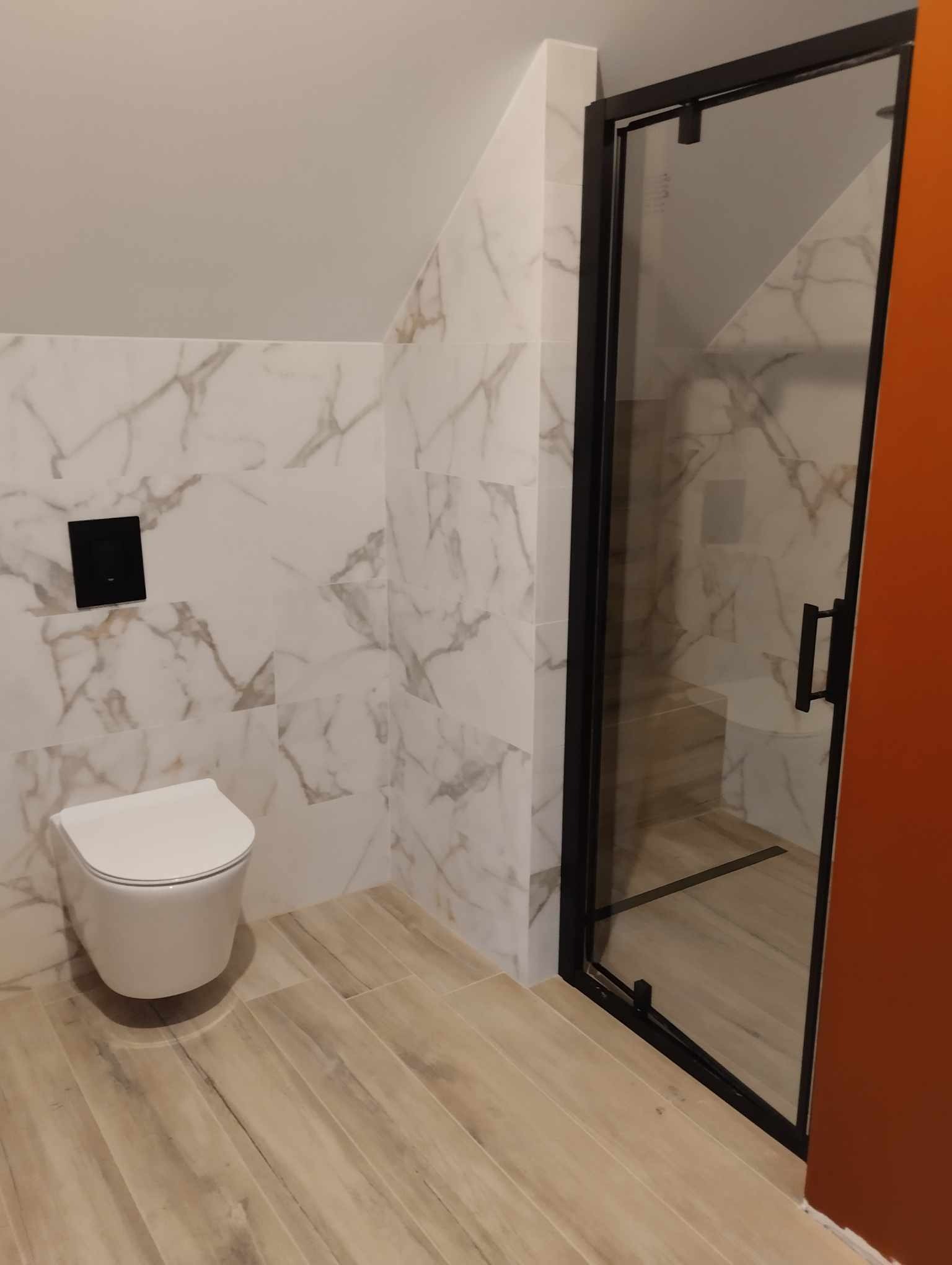 Wykończona łazienka na poddaszu: białe marmurowe płytki, podwieszana toaleta z czarnym przyciskiem, kabina prysznicowa z czarną ramą i drewnopodobna podłoga.