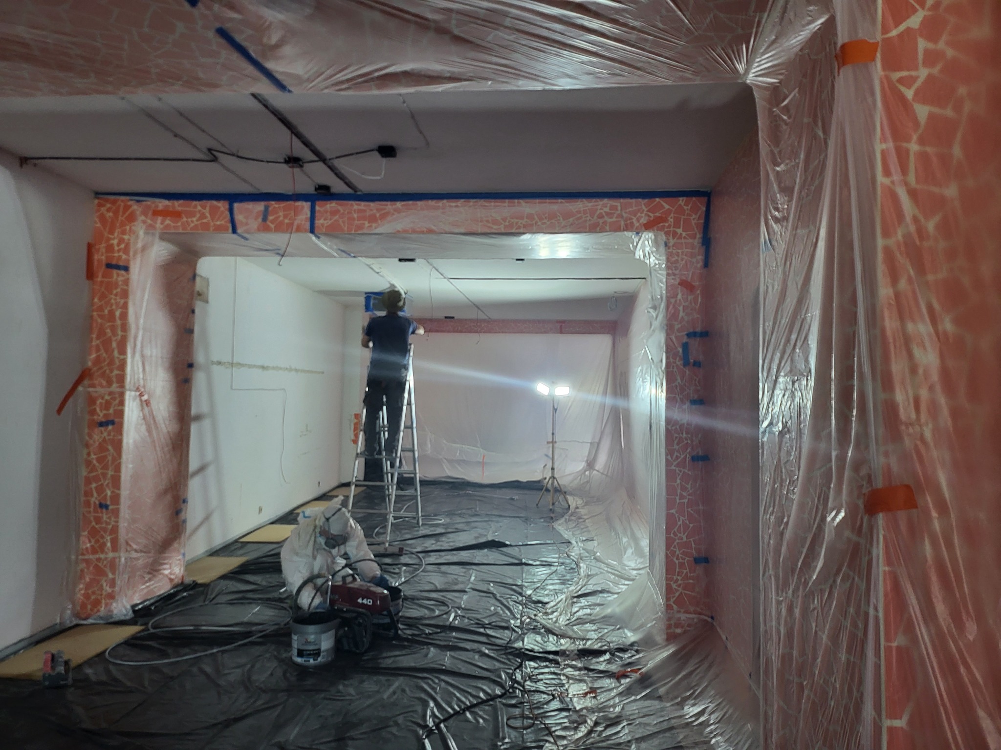 Remont pomieszczenia zabezpieczonego folią. Pracownik na drabinie, drugi w kombinezonie przy sprzęcie. Widoczne mozaiki na ścianach i suficie.