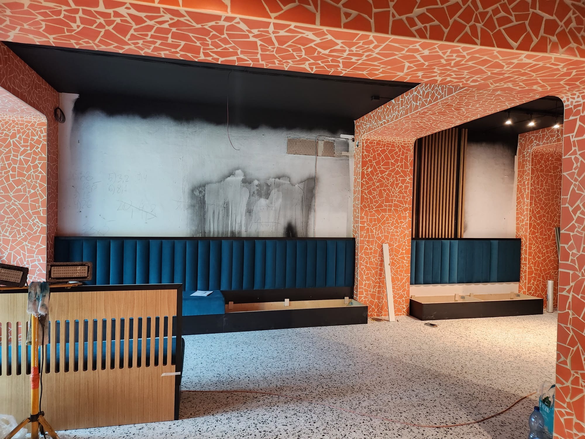 Wnętrze w trakcie wykończenia: mozaika na ścianach i suficie, zabudowa z niebieską tapicerką, surowa ściana z napisami, podłoga lastryko. Widoczne elementy montażowe.