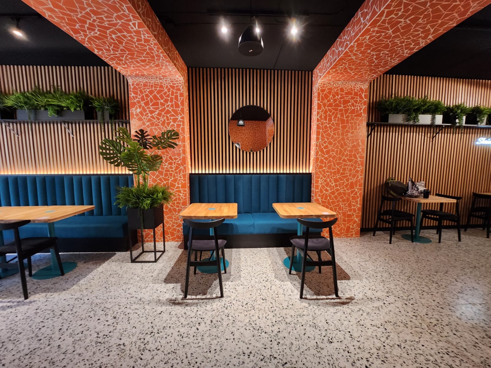Wykończone wnętrze lokalu: mozaika na kolumnach, drewniane lamele, turkusowa tapicerka sofy, stoliki z krzesłami i rośliny doniczkowe. Całość w nowoczesnym stylu.