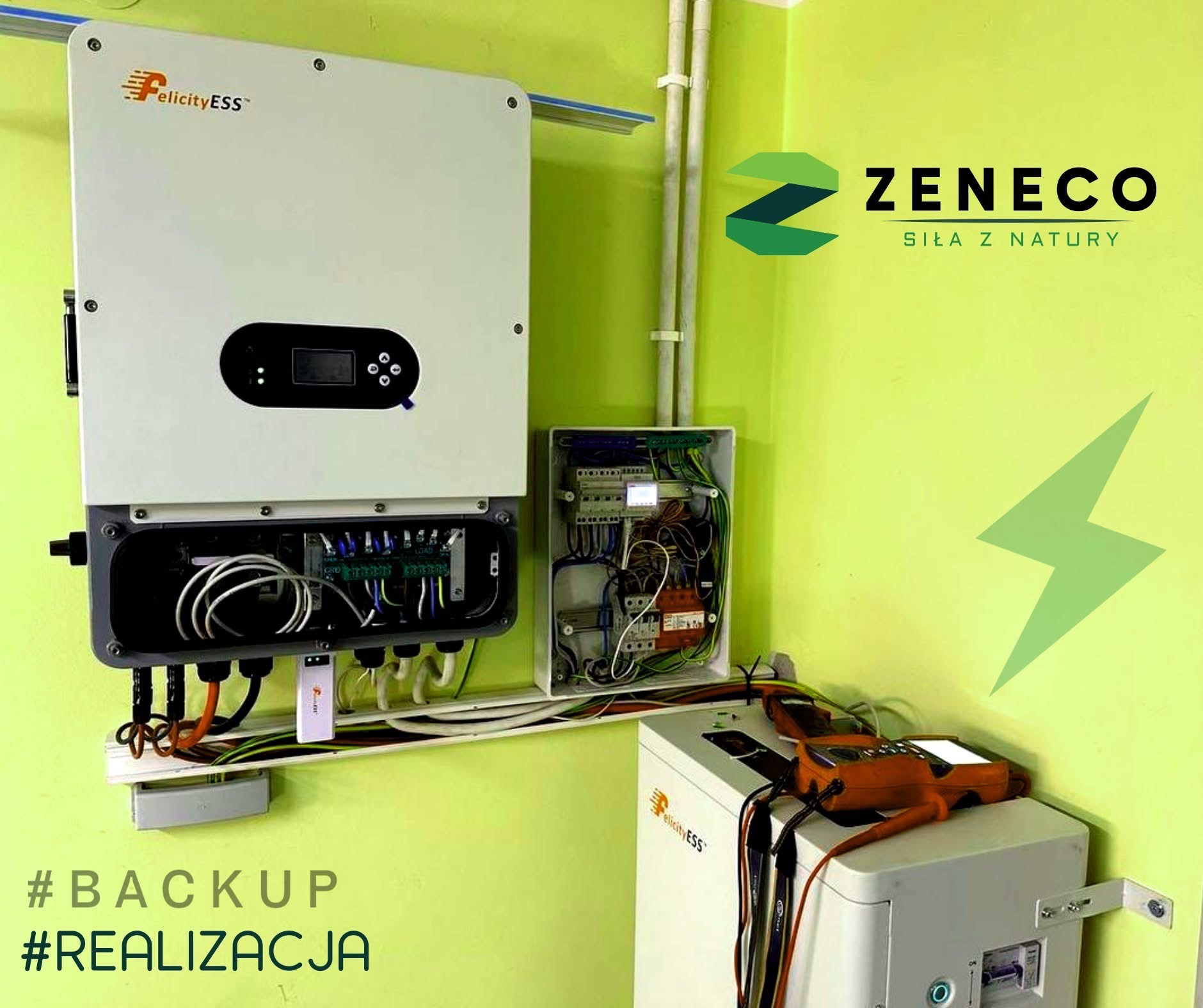 Instalacja magazynu energii Felicity ESS z inwerterem i rozdzielnicą na zielonej ścianie. Widoczne przewody, miernik i logo Zeneco.
