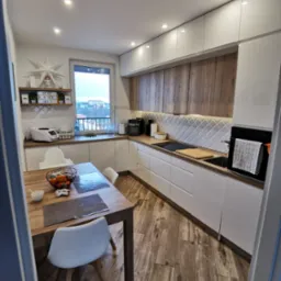 Nowoczesna kuchnia z białymi szafkami i drewnianymi akcentami. Widoczne oświetlenie podszafkowe, stół z owocami i krzesła. Aranżacja wnętrza z dbałością o detale.