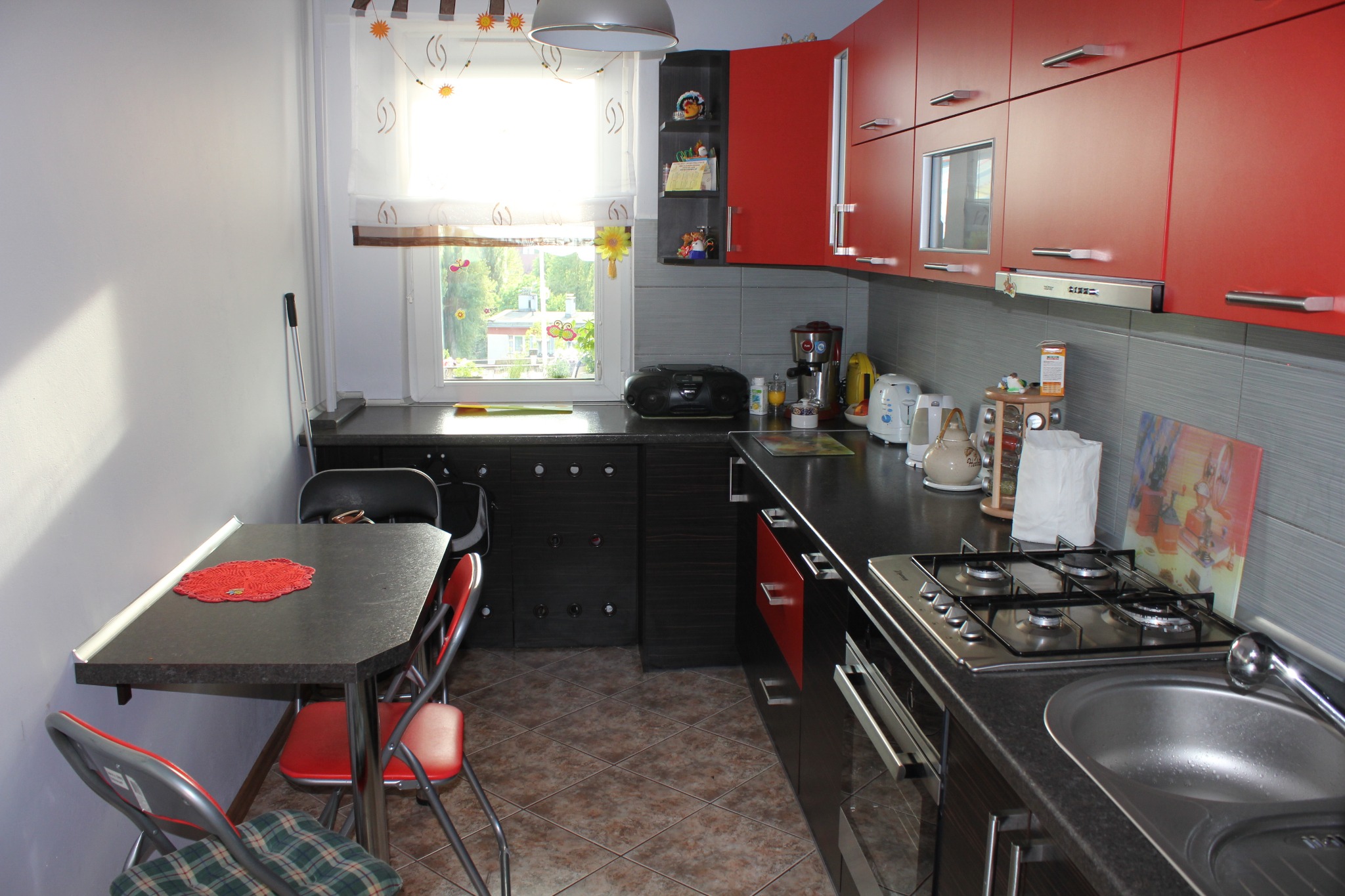 Kompaktowa kuchnia z czerwonymi szafkami górnymi i ciemnymi dolnymi, stół z krzesłami przy ścianie, okno z dekoracyjną roletą, brązowa podłoga.