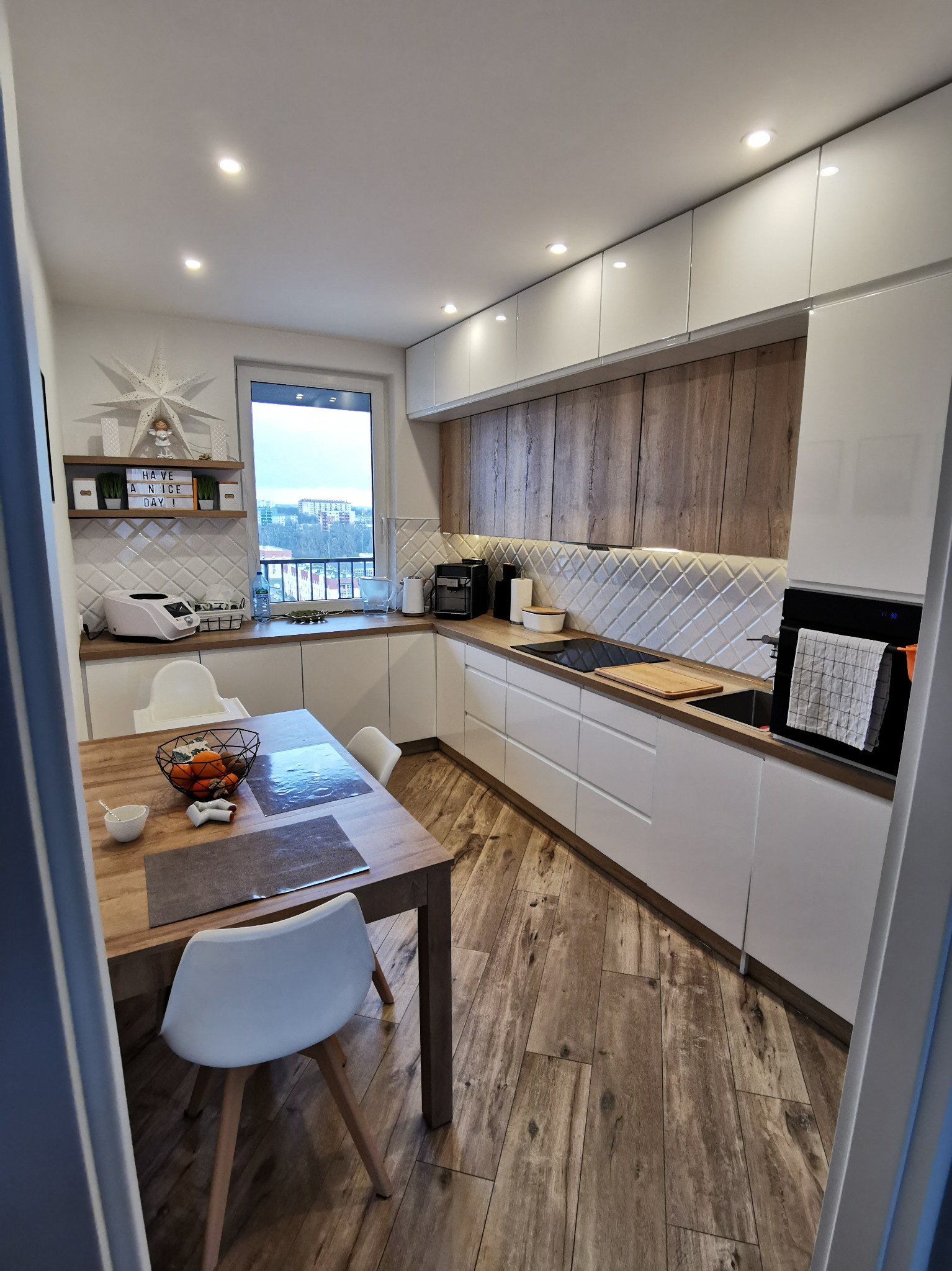 Nowoczesna kuchnia z białymi szafkami i drewnianymi akcentami. Widoczne oświetlenie podszafkowe, stół z owocami i krzesła. Aranżacja wnętrza z dbałością o detale.