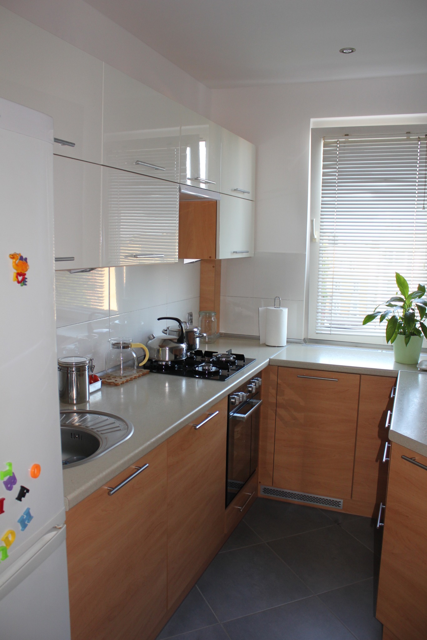 Jasna kuchnia z drewnianymi frontami szafek dolnych i białymi górnymi, okno z żaluzjami, czarny blat kuchenny z garnkami i płytą grzewczą.