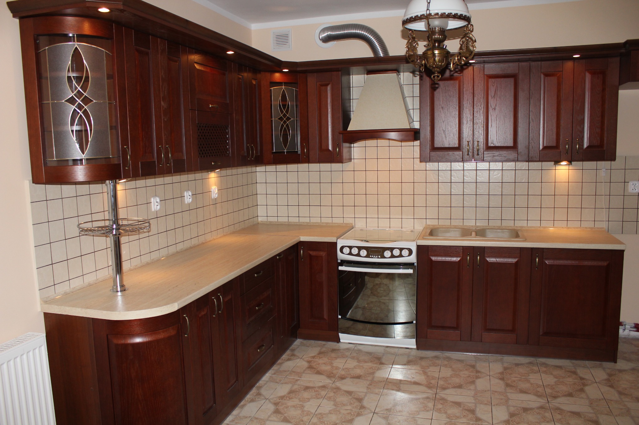 Ciemnobrązowa, klasyczna zabudowa kuchenna z przeszkloną szafką narożną, beżowym blatem i kremowymi płytkami. Widoczny fragment ozdobnego żyrandola.