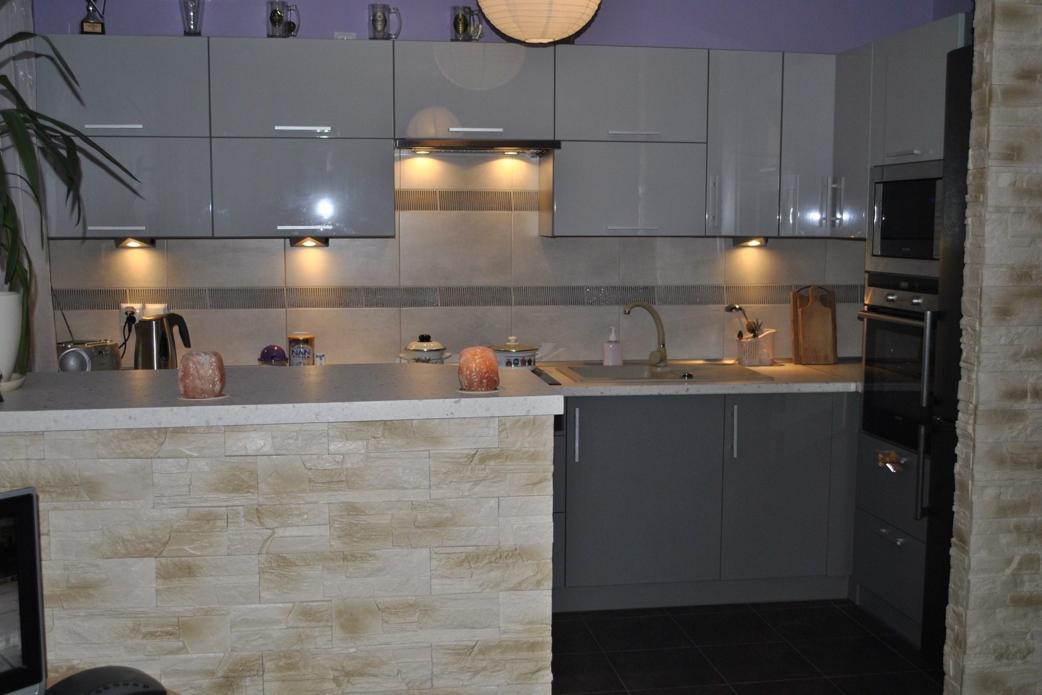 Szara zabudowa kuchenna z oświetleniem punktowym i blatem z dekoracyjnym kamieniem. Widoczny zlewozmywak, płyta grzewcza i sprzęt AGD w zabudowie.