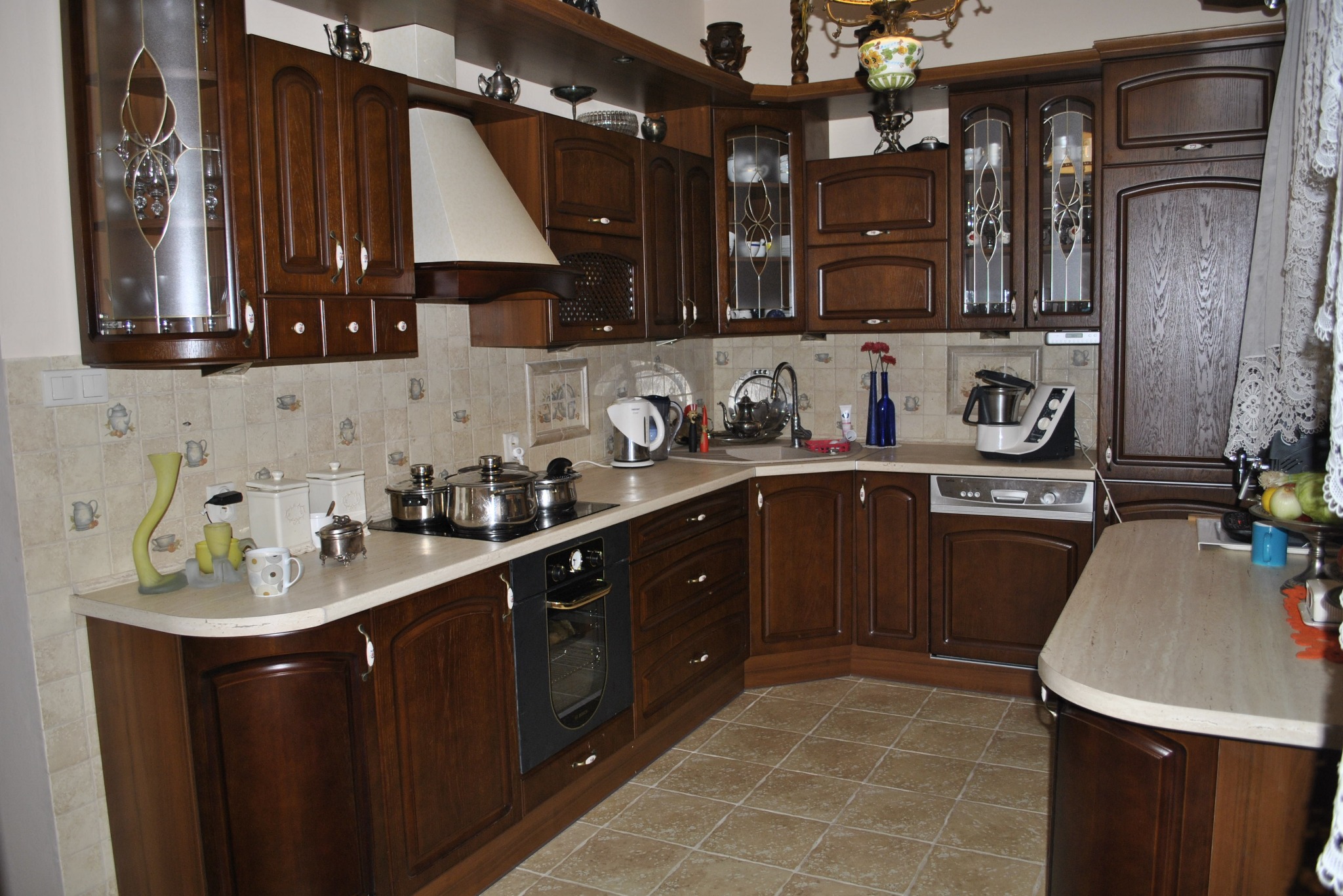 Klasyczna, brązowa kuchnia z frezowanymi frontami, szklanymi witrynami i beżowym blatem. Uwagę zwracają dekoracyjne akcesoria i płytki w tle.