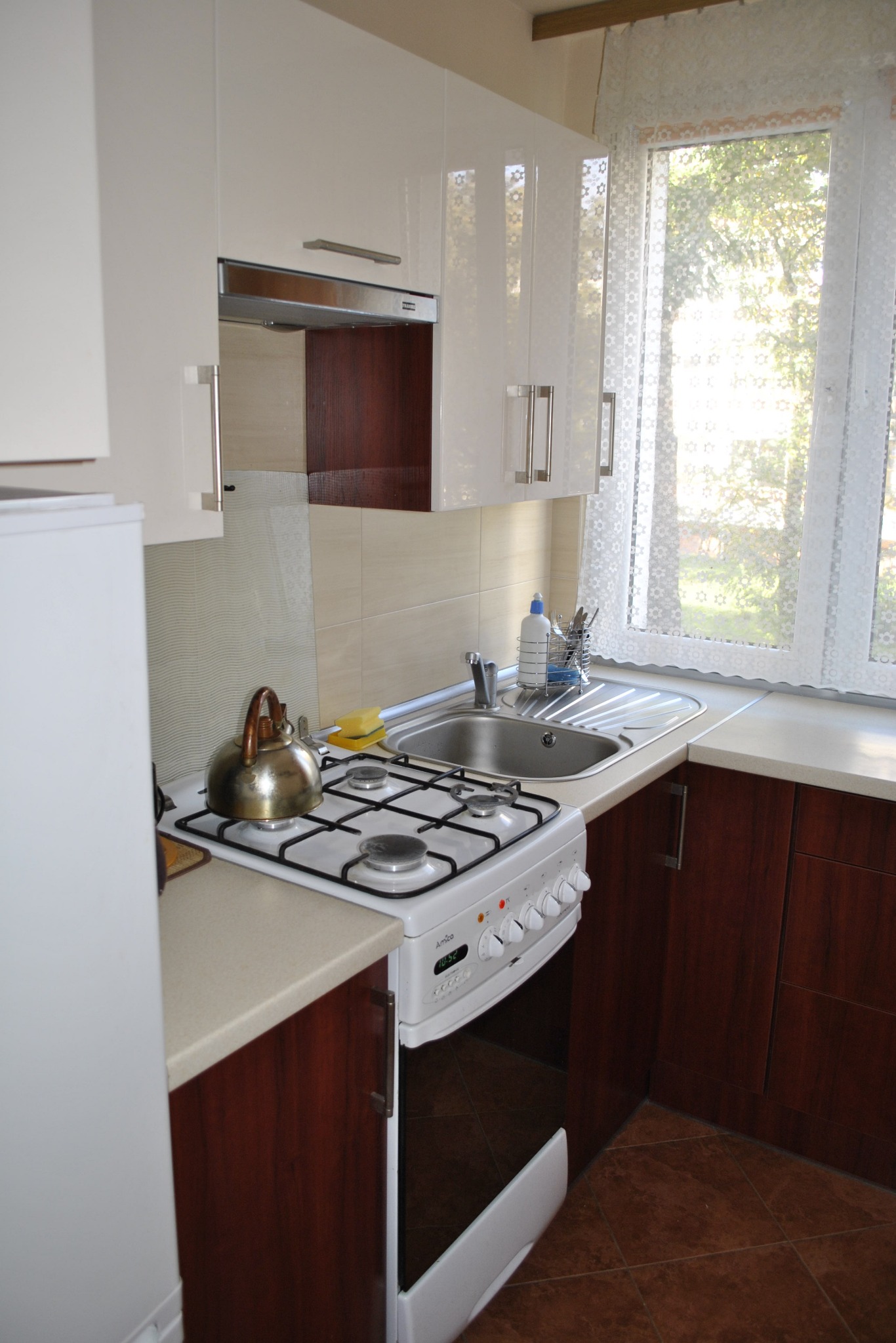 Jasna kuchnia z białymi szafkami i brązowymi akcentami. Widoczny zlew, kuchenka z czajnikiem i okno z firanką w tle, tworzące przytulną atmosferę.