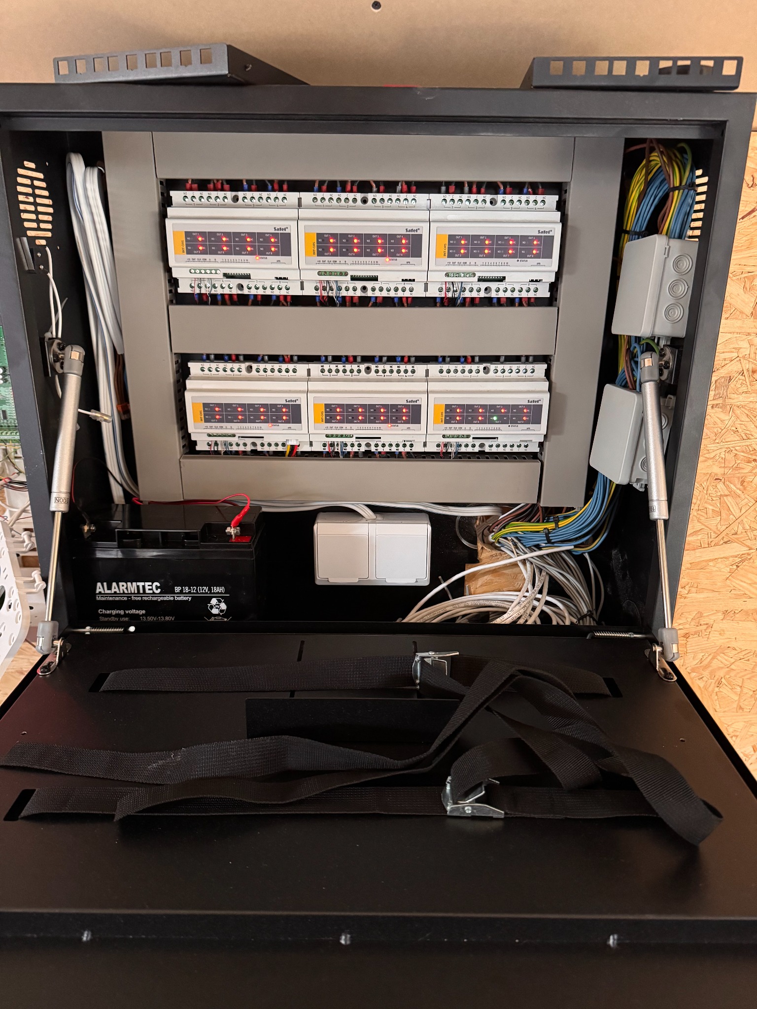 Otwarta szafka sterownicza z modułami alarmowymi Satel, akumulatorem Alarmtec i okablowaniem. Widoczne złącza, przewody i elementy montażowe.