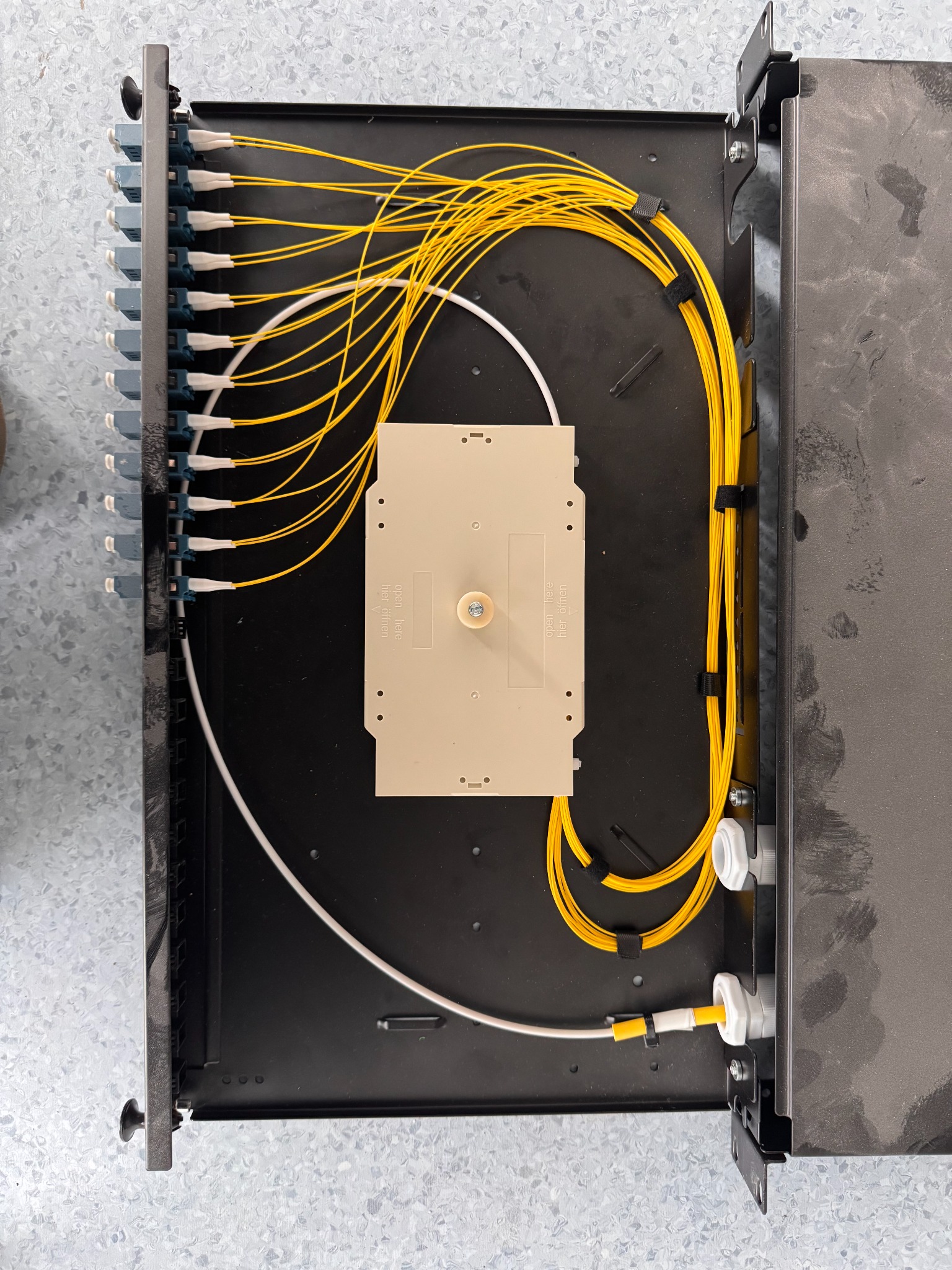 Rozdzielnica światłowodowa z żółtymi i białymi kablami, widok z góry. Kable uporządkowane wewnątrz czarnej obudowy. Element instalacji telekomunikacyjnej.