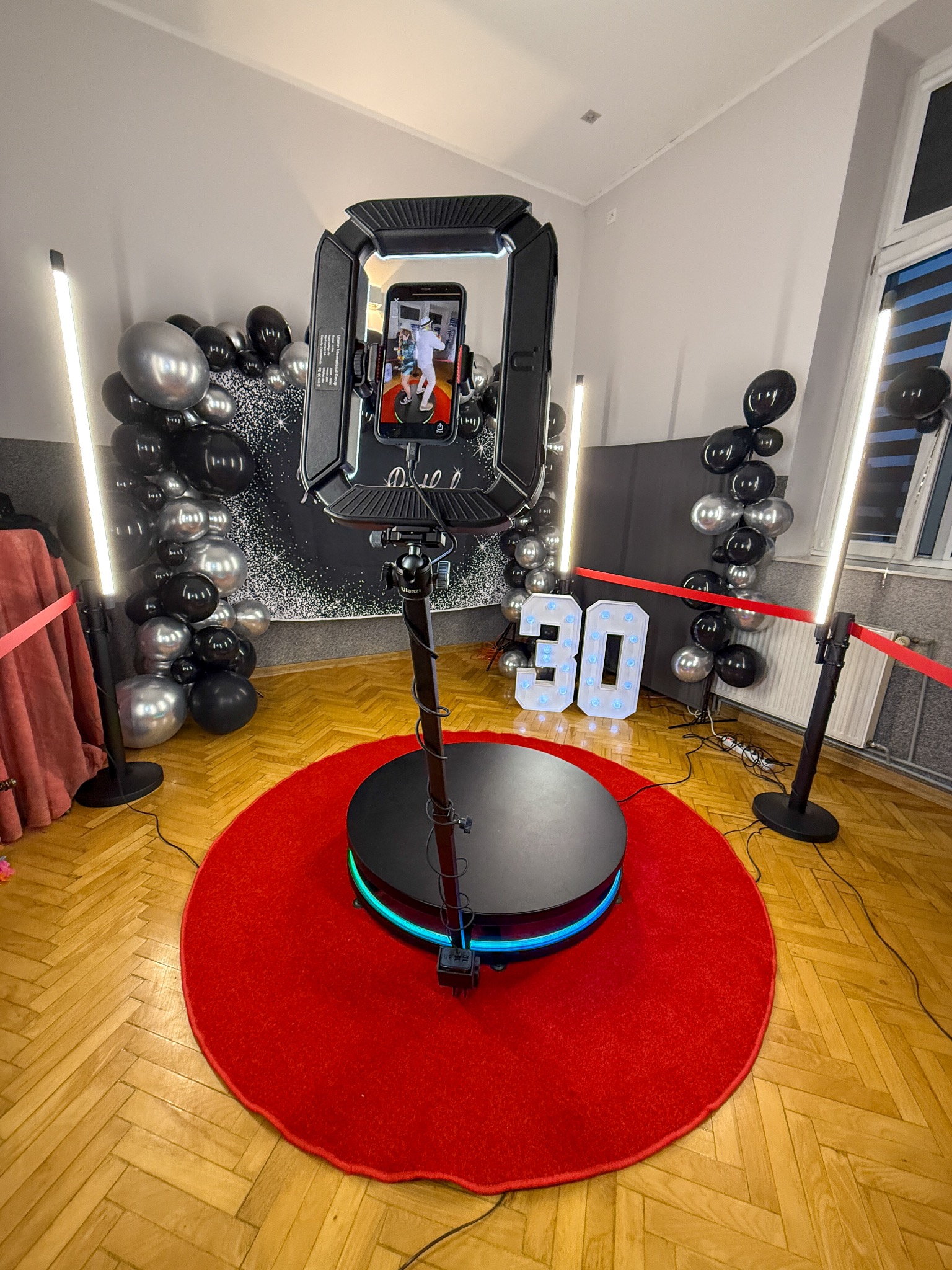 Oświetlona fotobudka 360 z platformą na czerwonym dywanie, dekoracje z balonów w odcieniach szarości i czerni, podświetlany numer 30, gotowa do nagrywania filmów.
