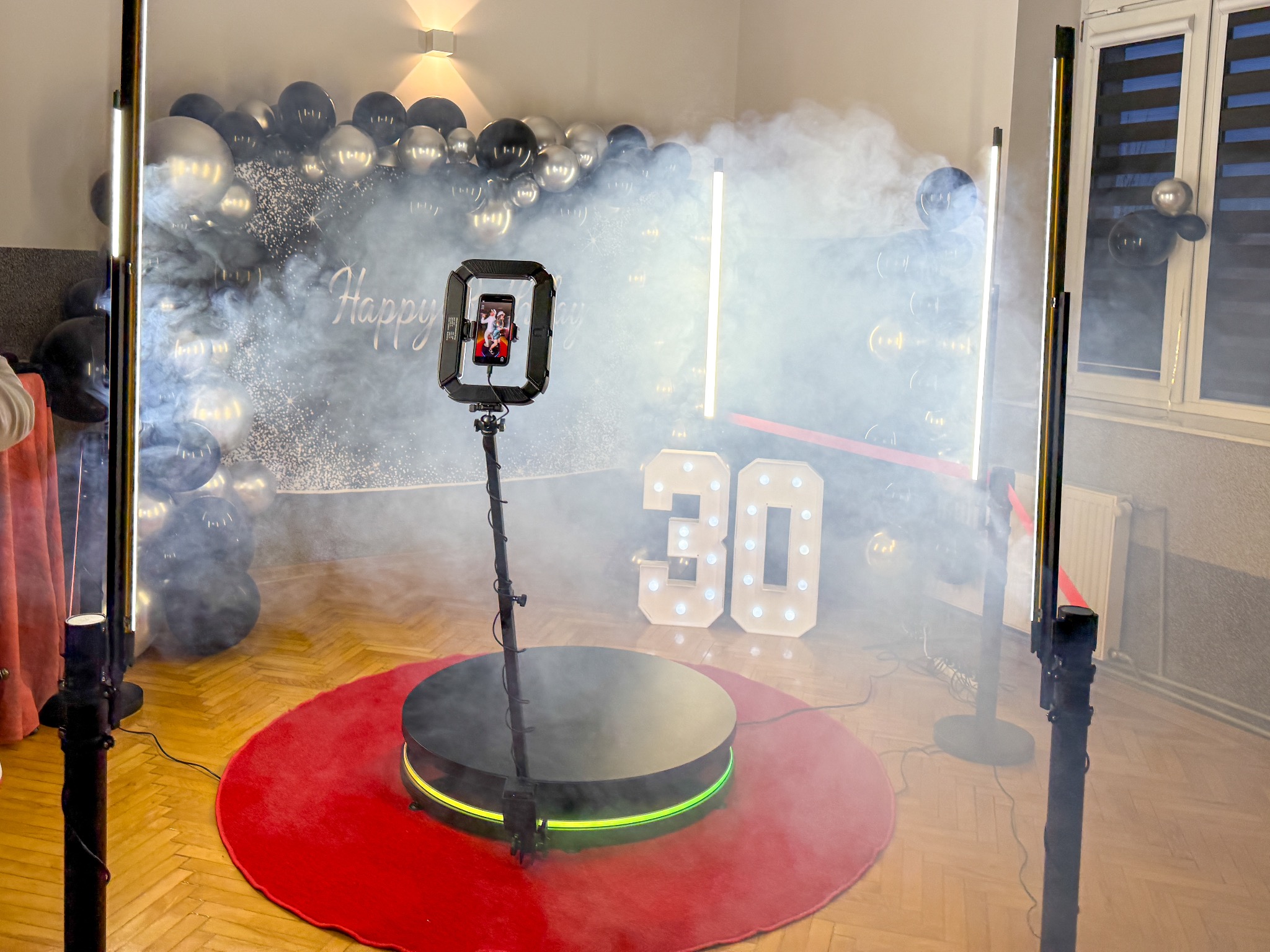 Rotacyjna fotobudka 360 na czerwonym dywanie, otoczona dekoracjami urodzinowymi i sztucznym dymem. Na ekranie widoczny uśmiechnięty mężczyzna w czerwonej koszuli.