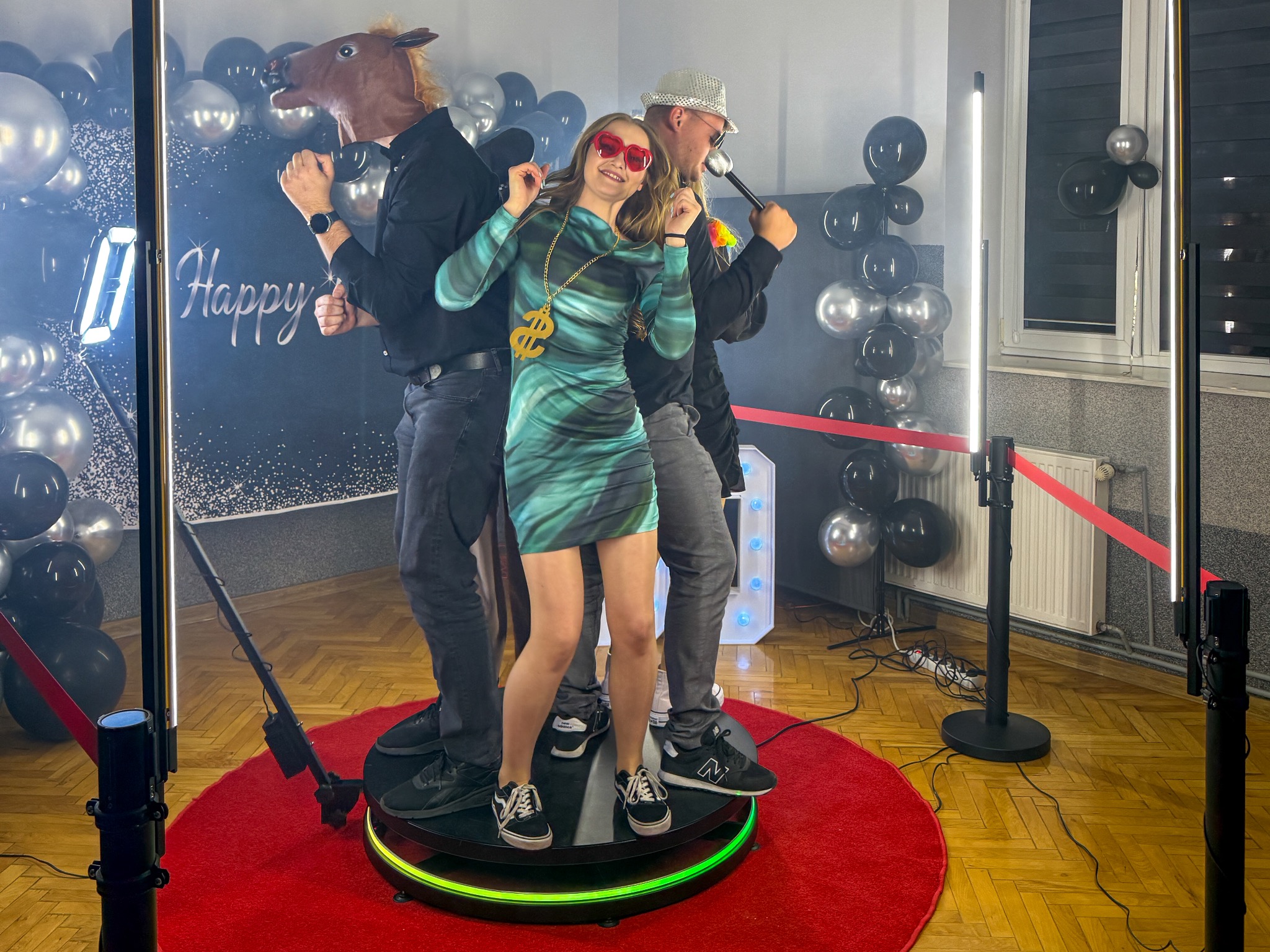Trzy osoby na platformie 360 fotobudki: kobieta w okularach i naszyjniku dolarowym, mężczyzna w masce konia i mężczyzna w kapeluszu, czarne i srebrne balony w tle.