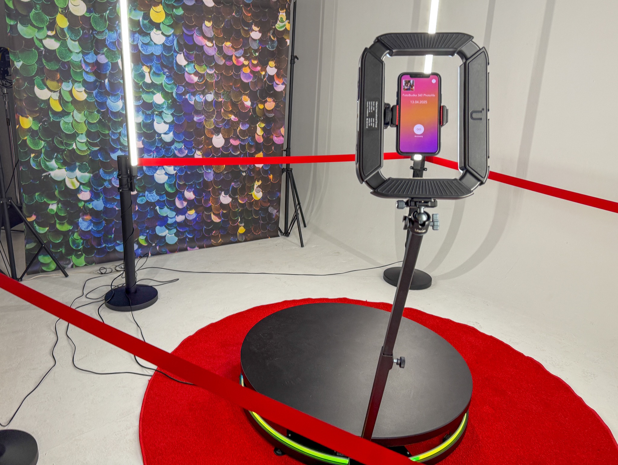 Profesjonalna fotobudka 360 z oświetleniem LED, tłem z cekinami i czerwonym dywanem, gotowa do użycia na imprezie. Widoczny ekran smartfona z interfejsem aplikacji.