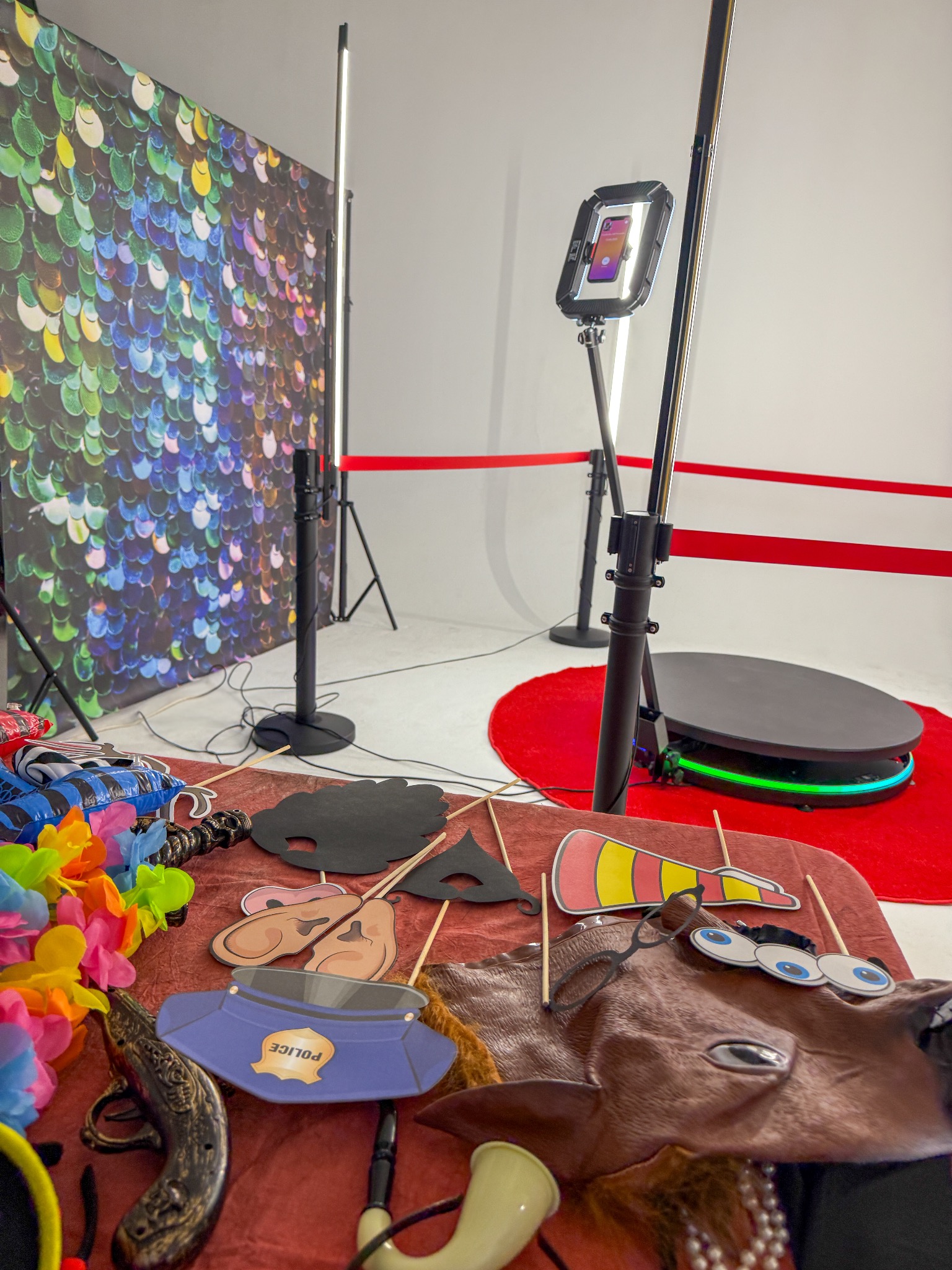 Stanowisko fotobudki 360 z rekwizytami: maski, okulary, kapelusz policjanta. W tle kolorowa ścianka, lampy i platforma obrotowa z czerwonym dywanem.