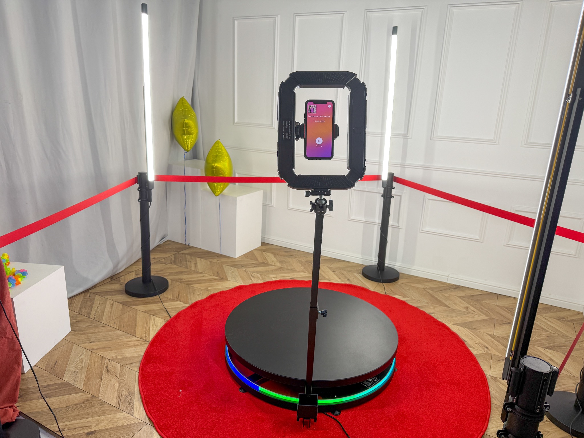 Obracana platforma 360° z uchwytem na smartfon i oświetleniem LED na czerwonym dywanie, otoczona słupkami z taśmą. W tle białe panele ścienne i złote balony.