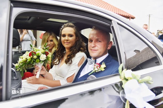 Uśmiechnięta para młoda w samochodzie, panna młoda trzyma bukiet białych i czerwonych róż, w tle druhny i orkiestra. Ślubna sesja w plenerze.