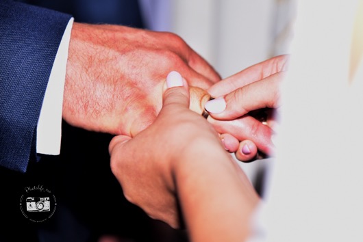Zbliżenie dłoni podczas ceremonii ślubnej, panna młoda zakłada obrączkę na palec pana młodego. Widoczny fragment granatowej marynarki.