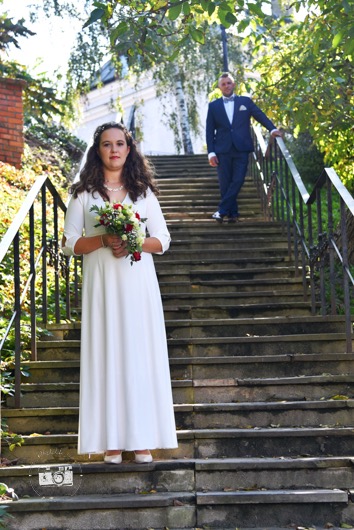 Panna młoda w białej sukni z bukietem na schodach, pan młody w granatowym garniturze w tle. Ujęcie w plenerze, w słoneczny dzień.