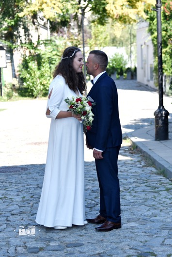 Para młoda wpatrzona w siebie na brukowanej uliczce, panna młoda w białej sukni z bukietem, pan młody w granatowym garniturze, w tle zieleń i budynki.