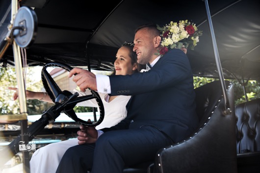 Uśmiechnięta para młoda w zabytkowym samochodzie z bukietem kwiatów. Pan młody w ciemnym garniturze, panna młoda w białej sukni. Kadr z sesji ślubnej w plenerze.