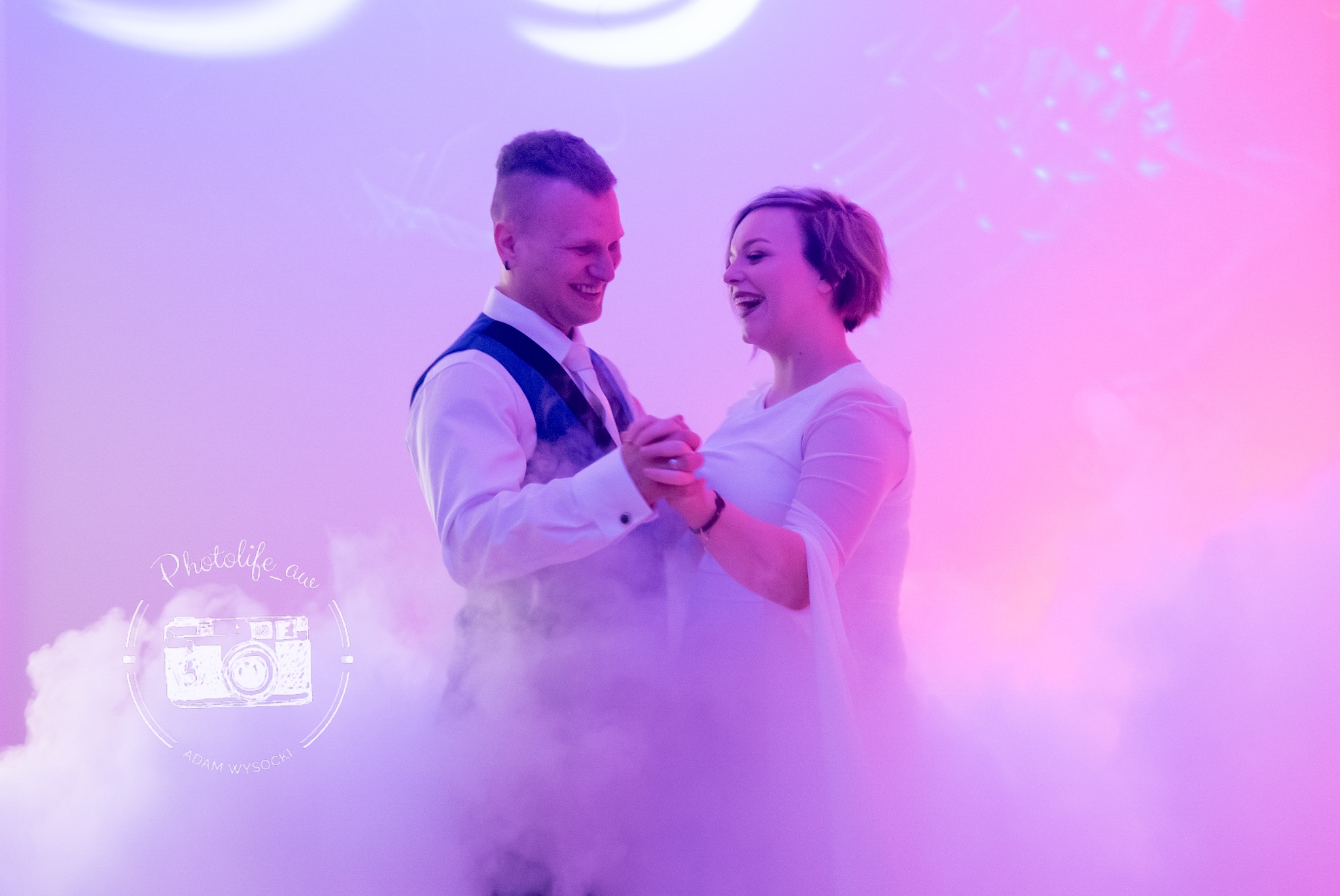 Uśmiechnięta para młoda w tańcu, otulona mgłą na fioletowo-różowym tle. Widoczny logo fotografa w lewym dolnym rogu.